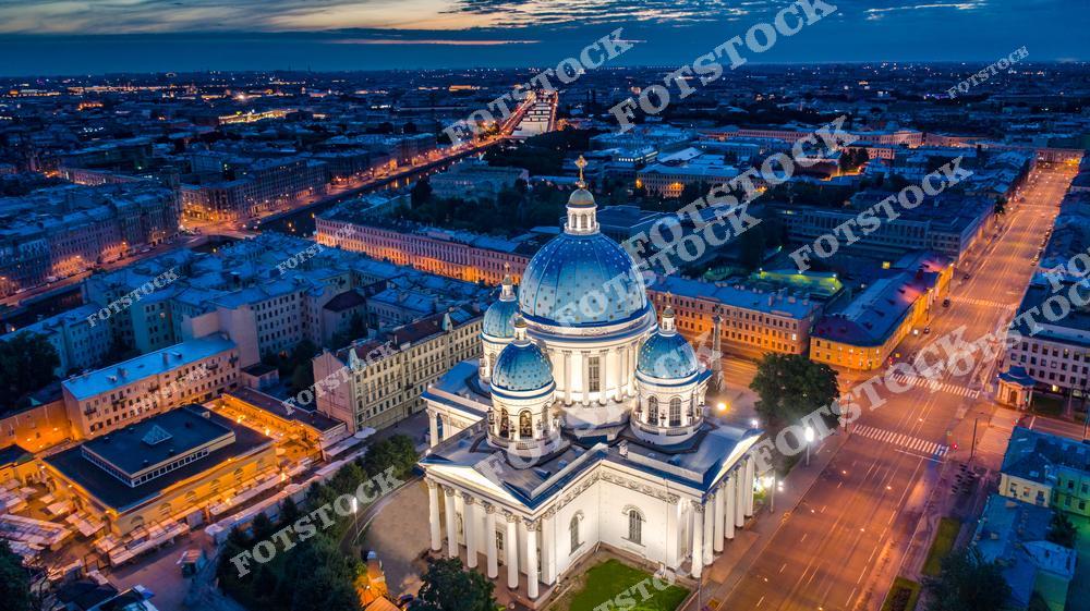 Панорамная фотография вечернего Санкт-Петербурга, снятая с дрона с высоты птичьего полета. На снимке запечатлен Троице-Измайловский собор с голубыми куполами, освещенные улицы исторического центра и архитектура ночного Петербурга. Городские огни создают атмосферу уютного вечернего мегаполиса, а высотный ракурс позволяет увидеть масштаб и красоту Северной столицы России. Фотография передает уникальный стиль архитектуры Санкт-Петербурга, широкие проспекты и знаменитые достопримечательности города. Снимок идеально подходит для туристических проектов, публикаций о путешествиях, рекламы, оформления интерьеров и фотостоков с видами Петербурга.
